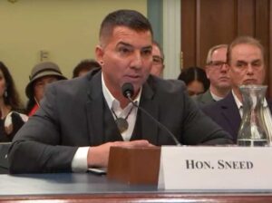 Photo of Chief of Eastern Band of Cherokee Indians, Sneed, from U.S. Congressional office official photos. He is wearing bow-low, white shirt and suit jacket at table with white sign in front of him with words "Hon. Sneed". People sit in background